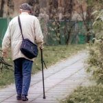 Hombre mayor caminando con confianza por una acera urbana arbolada utilizando un bastón ergonómico plegable contemporáneo durante el atardecer.