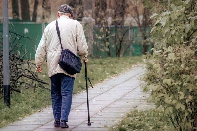 Hombre mayor caminando con confianza por una acera urbana arbolada utilizando un bastón ergonómico plegable contemporáneo durante el atardecer.